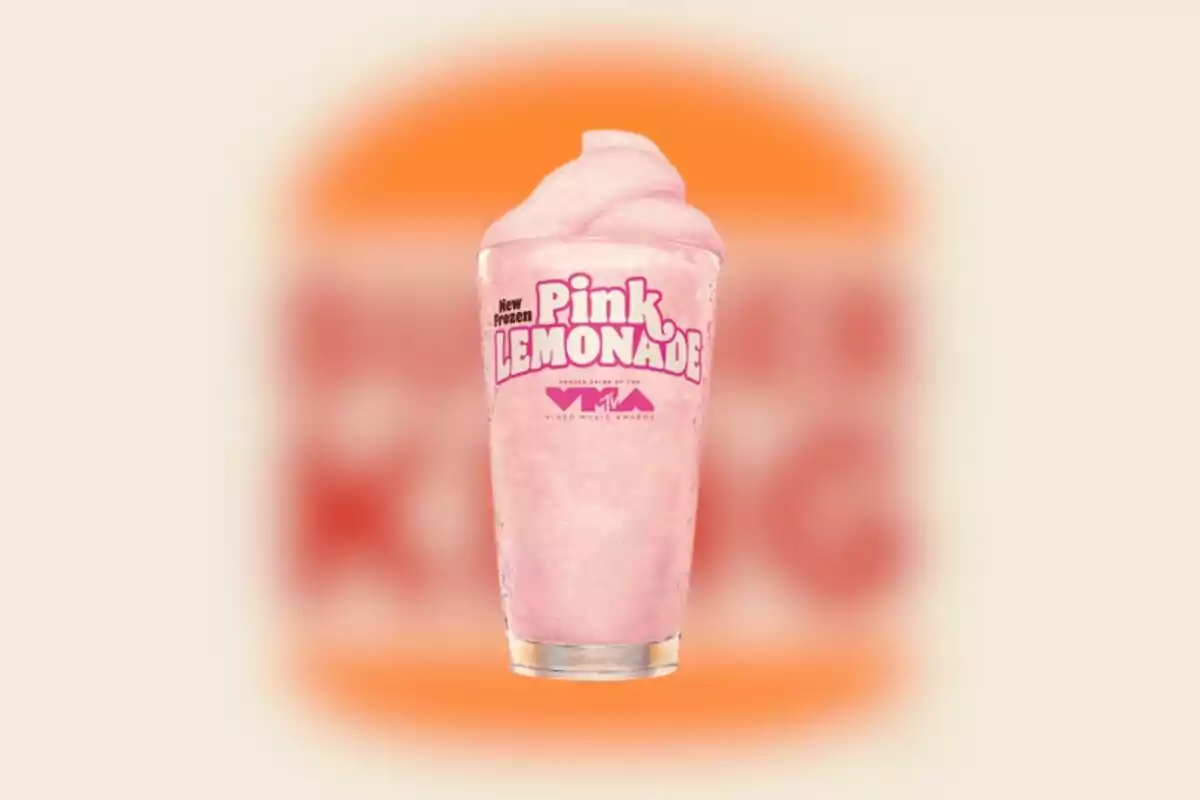 Transparent cup with pink lemonade frappé and Pink Lemonade logo on the front, set against a blurred orange and beige background Transparent cup with pink lemonade frappé and Pink Lemonade logo on the front, set against a blurred orange and beige background