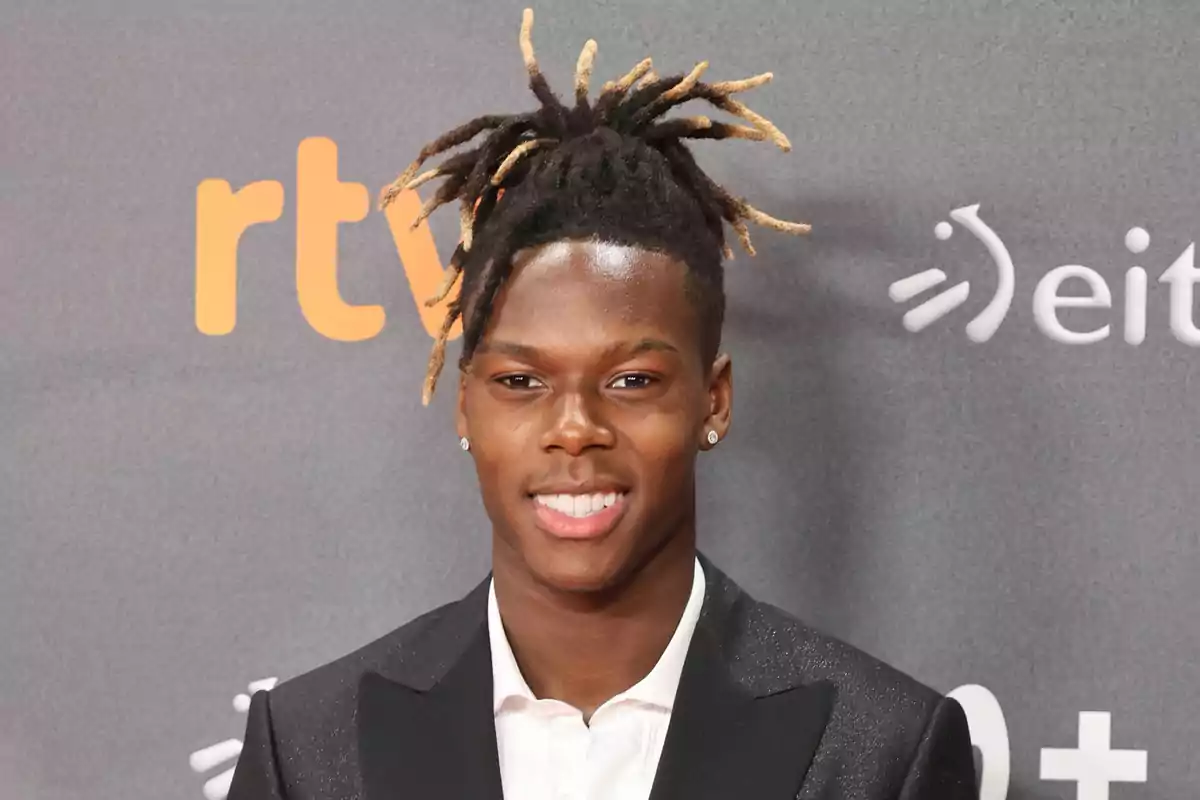 Nico Williams amb vestit fosc i camisa blanca somrient davant d’un fons gris amb logotips.