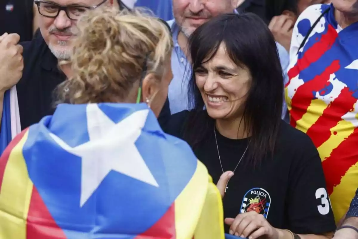 Dona somrient i conversant amb una altra persona que porta una bandera estelada sobre les espatlles en una reunió o manifestació envoltades de més persones Dona somrient i conversant amb una altra persona que porta una bandera estelada sobre les espatlles en una reunió o manifestació envoltades de més persones