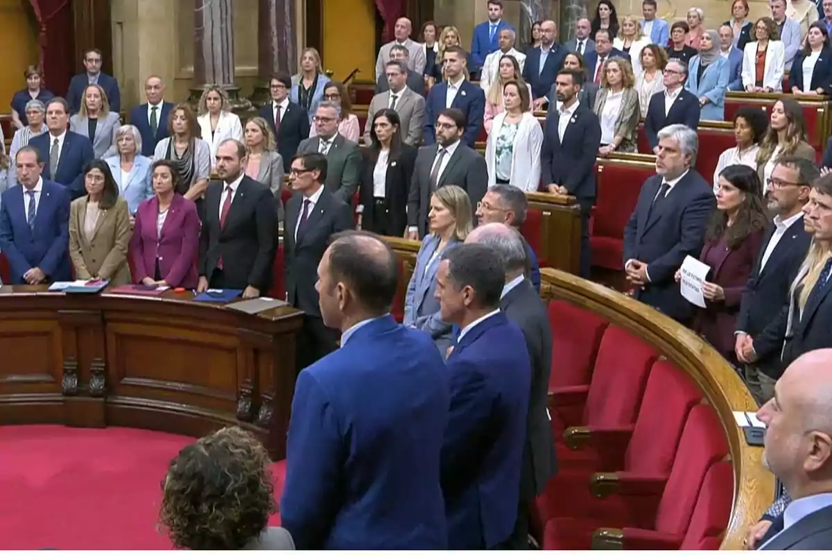 Un grupo numeroso de personas de pie en un recinto parlamentario con asientos rojos y ambiente formal