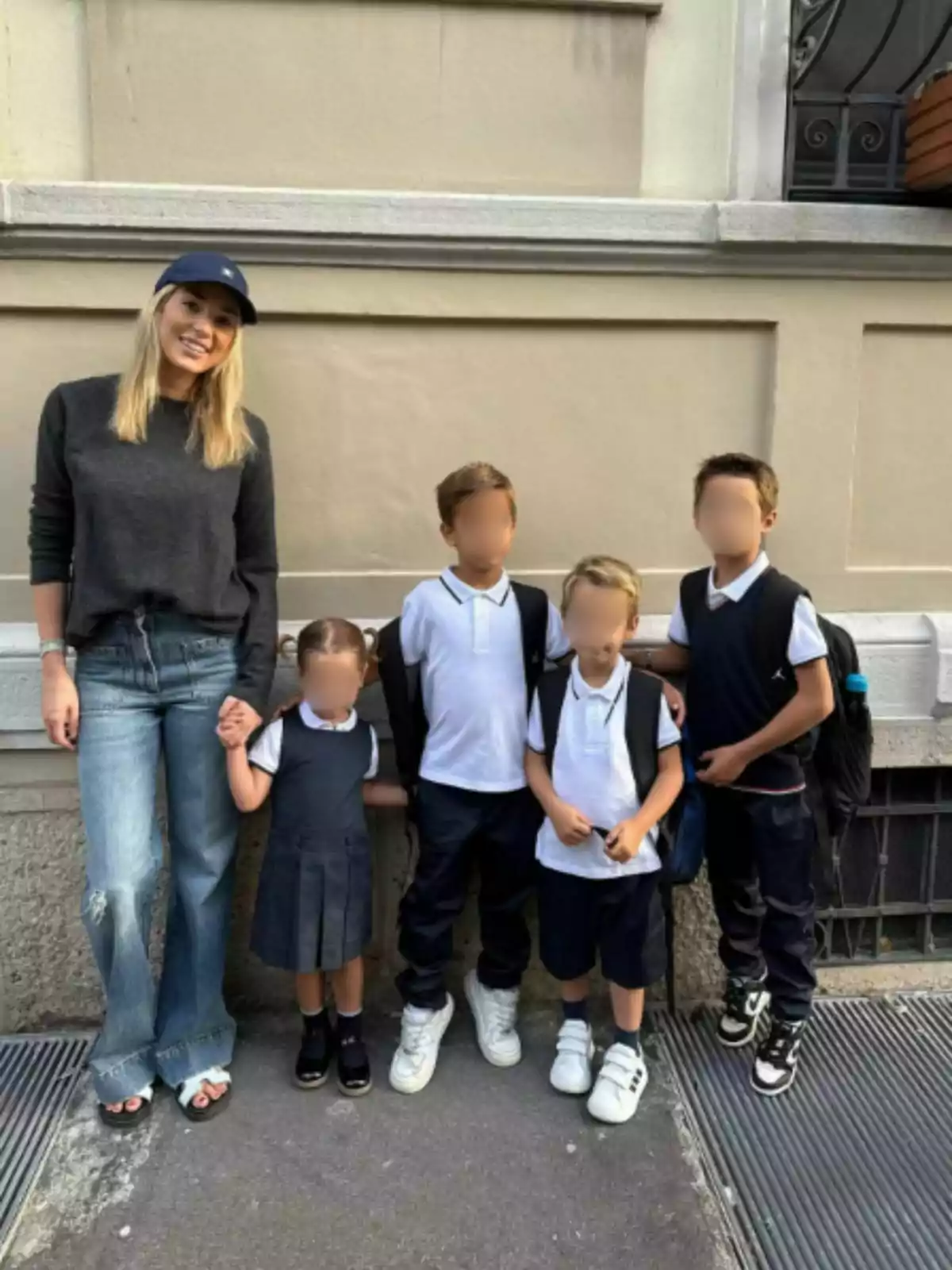 Alice Campello amb gorra i jersei fosc sosté la mà d’una nena petita amb uniforme escolar mentre posen al costat de tres nens també vestits amb uniforme davant d’una paret beix. Alice Campello amb gorra i jersei fosc sosté la mà d’una nena petita amb uniforme escolar mentre posen al costat de tres nens també vestits amb uniforme davant d’una paret beix.