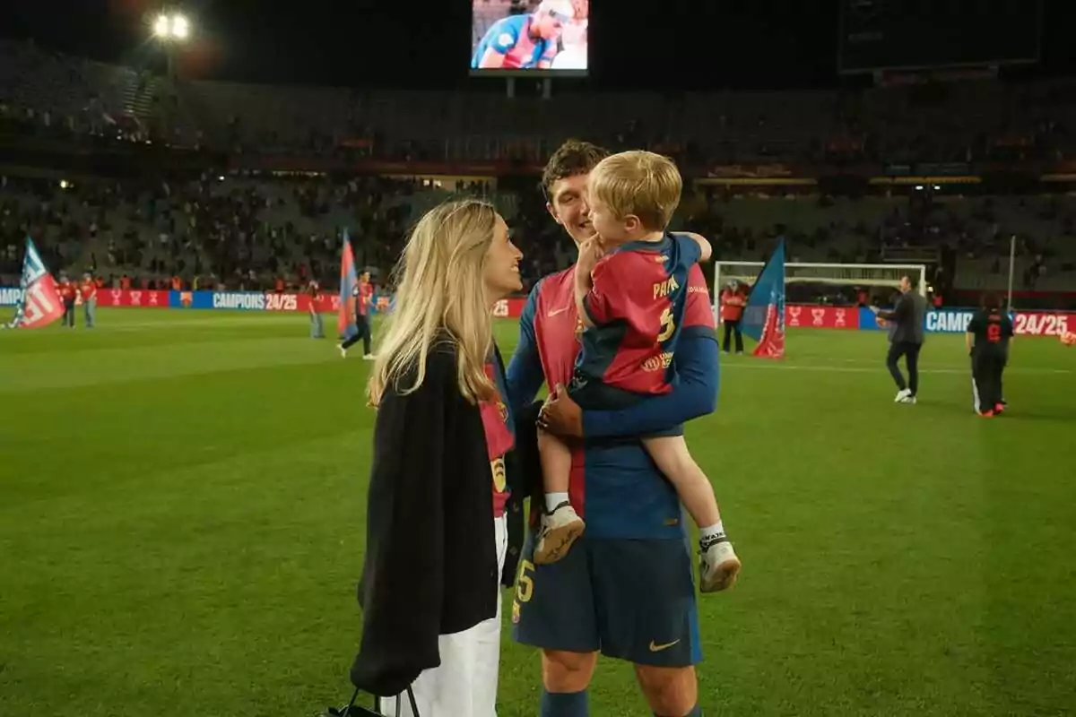 Christensen amb l’uniforme blau i vermell sosté un nen petit en braços mentre conversa somrient amb Katrine Friis al camp d’un estadi després d’un partit.