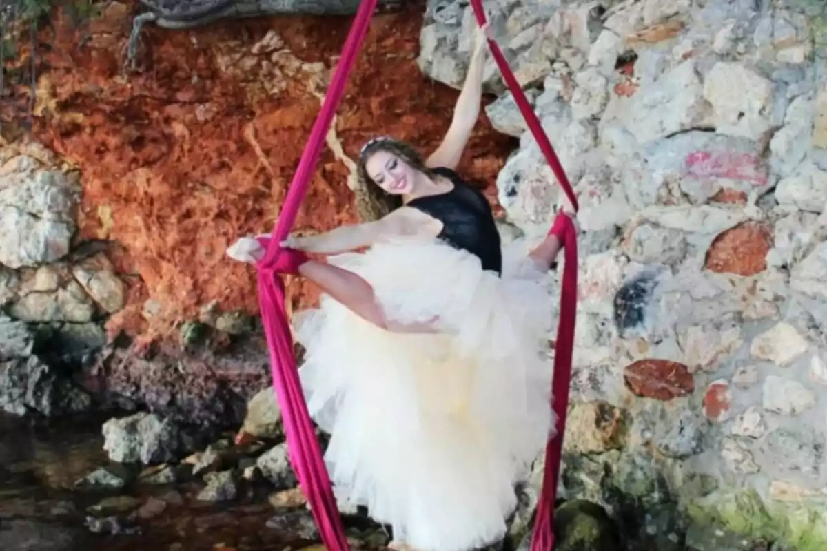 Ballarina amb tutú blanc i mallot negre fent acrobàcies en teles vermelles davant d’una paret de pedra