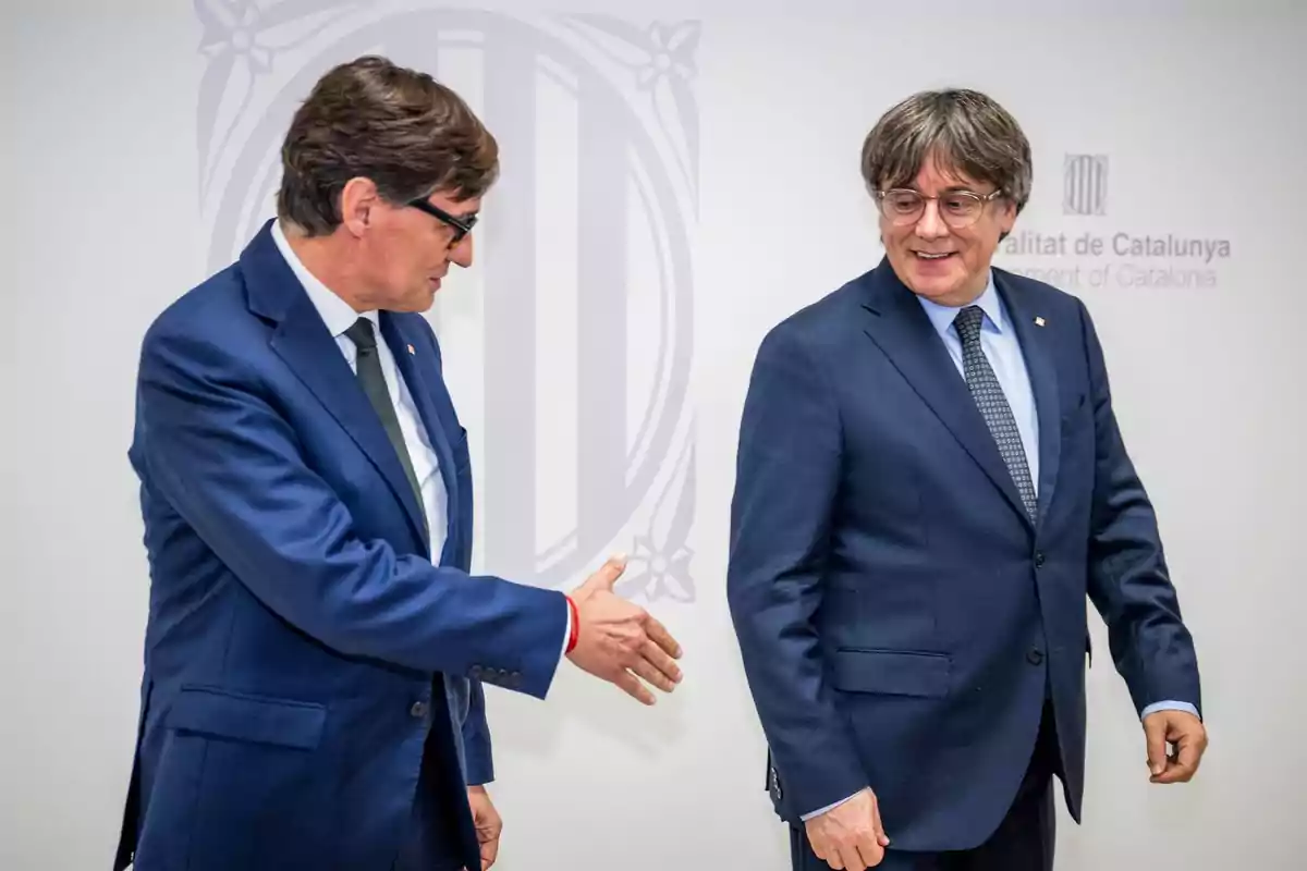 Salvador Illa i Carles Puigdemont amb vestit blau es troben en un entorn institucional, un d’ells allarga la mà per saludar mentre l’altre somriu i mira cap a un costat. Salvador Illa i Carles Puigdemont amb vestit blau es troben en un entorn institucional, un d’ells allarga la mà per saludar mentre l’altre somriu i mira cap a un costat.