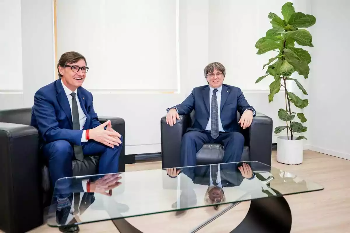 Carles Puigdemont i Salvador Illa amb vestit blau asseguts en butaques negres conversen en una oficina moderna amb una taula de vidre i una planta gran al fons. Carles Puigdemont i Salvador Illa amb vestit blau asseguts en butaques negres conversen en una oficina moderna amb una taula de vidre i una planta gran al fons.