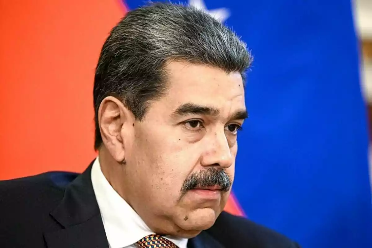 Primer pla de Nicolás Maduro amb bigoti i amb expressió seriosa, vestit amb vestit i corbata, davant d’un fons blau i vermell. Primer pla de Nicolás Maduro amb bigoti i amb expressió seriosa, vestit amb vestit i corbata, davant d’un fons blau i vermell.