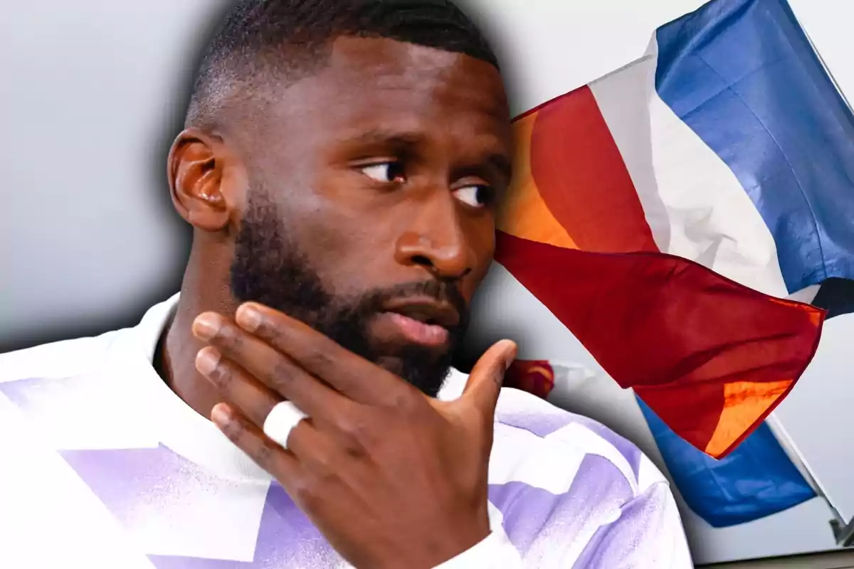 Home pensatiu amb barba i samarreta blanca i lila davant d’una bandera tricolor onejant