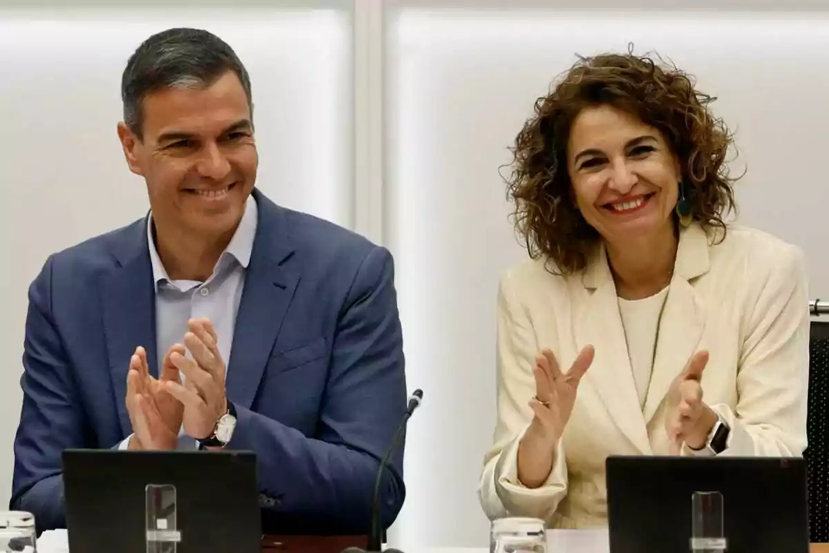 Dos personas sonrientes aplauden sentadas en una mesa de reuniones con portátiles frente a ellas