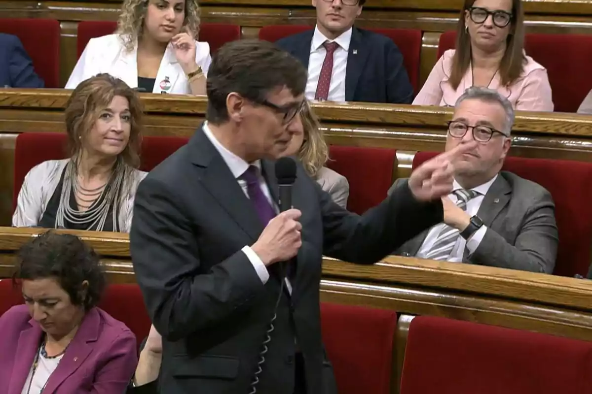 Un hombre de traje y gafas sostiene un micrófono mientras habla en una sala con asientos rojos rodeado de otras personas que lo observan atentamente