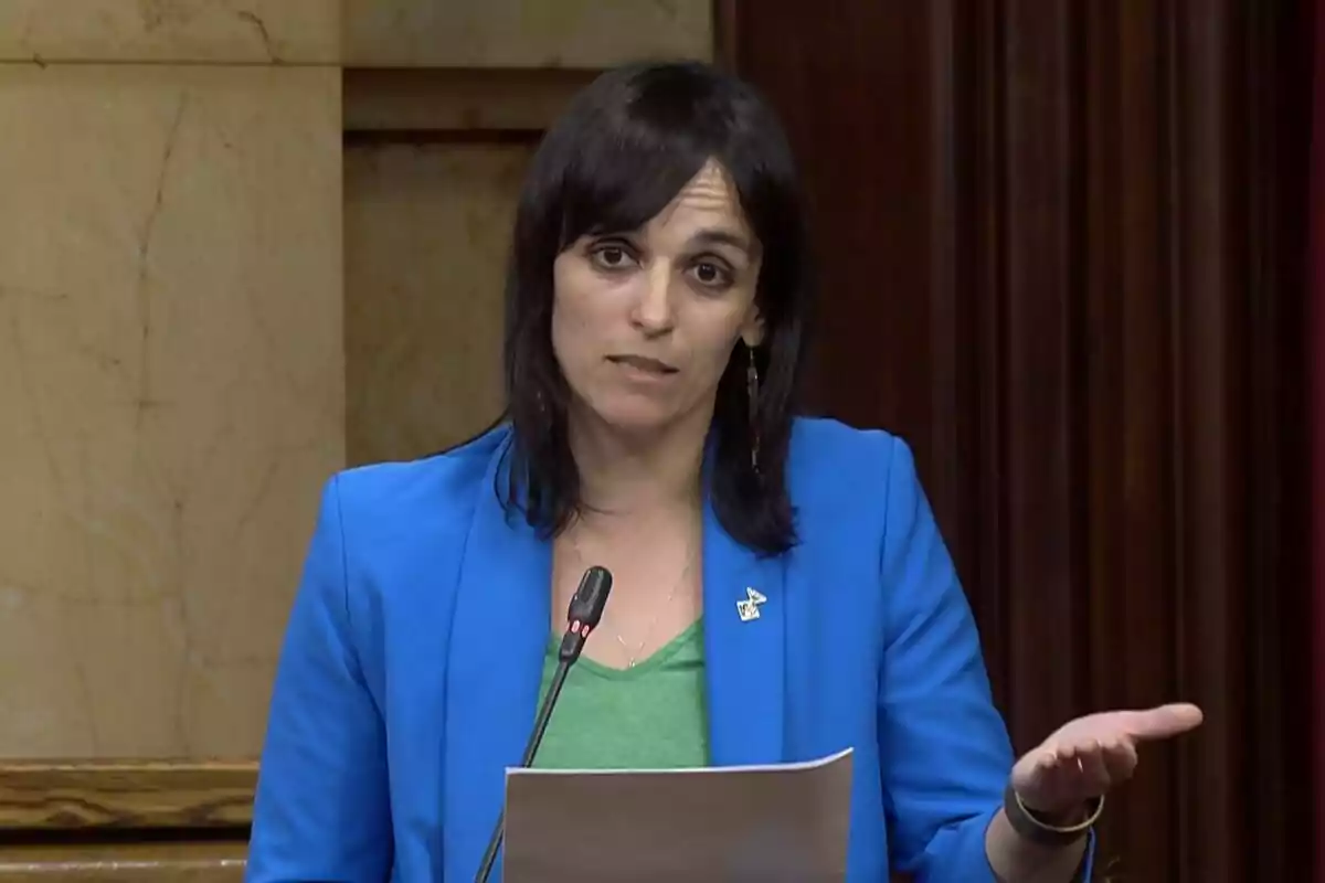 Mujer de cabello oscuro con chaqueta azul hablando frente a un micrófono y sosteniendo una hoja de papel