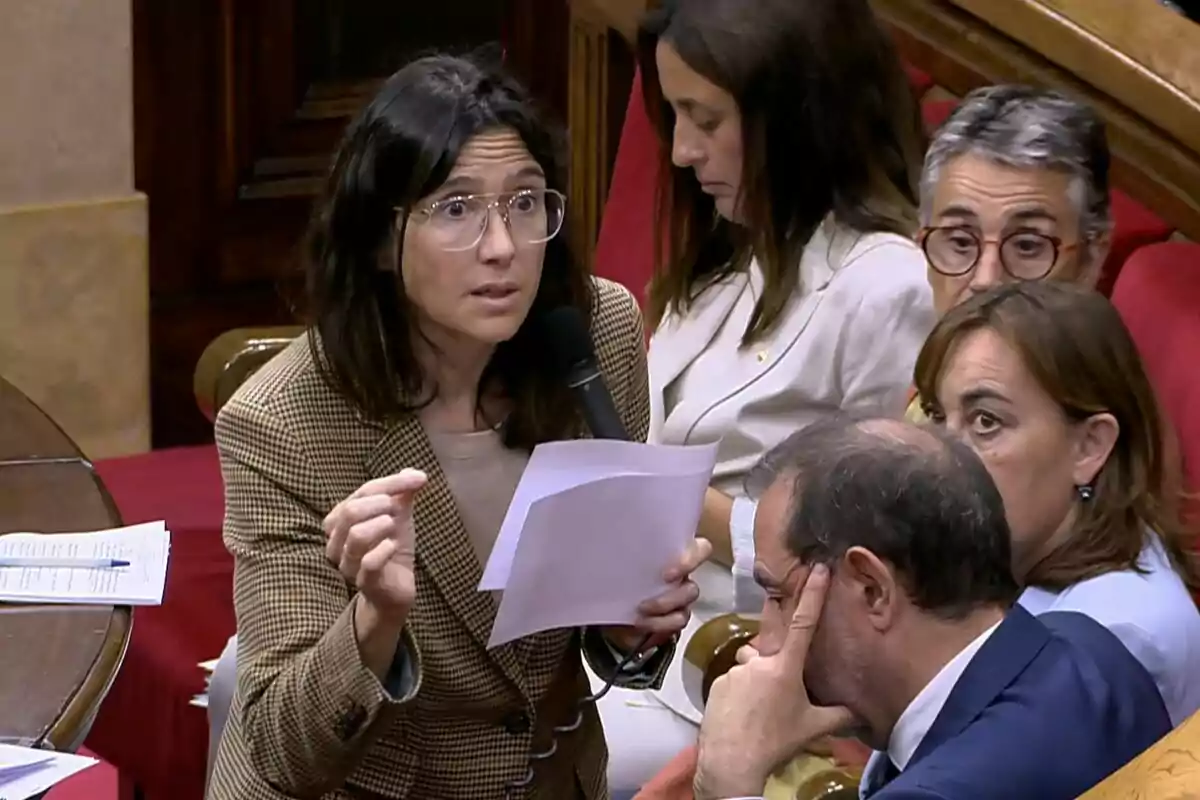 Mujer con gafas y chaqueta de cuadros hablando con un micrófono y sosteniendo papeles en una sala de reuniones rodeada de otras personas atentas