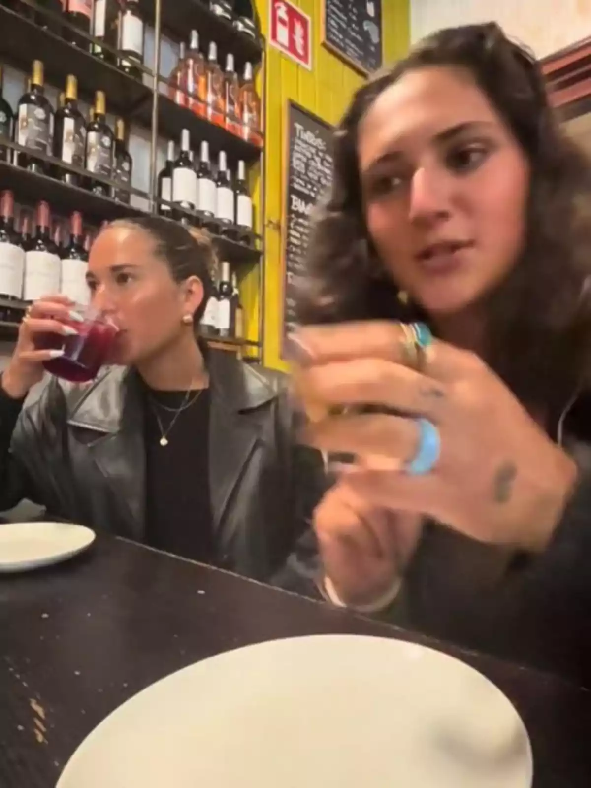 Dues dones estan assegudes en una taula de restaurant, una d’elles beu d’un got mentre l’altra parla i gesticula, al fons es veuen prestatges amb ampolles de vi. Dues dones estan assegudes en una taula de restaurant, una d’elles beu d’un got mentre l’altra parla i gesticula, al fons es veuen prestatges amb ampolles de vi.