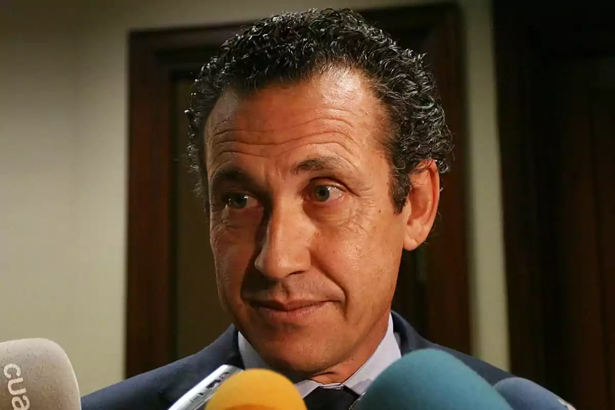 Home de cabell arrissat i vestit fosc responent preguntes davant de diversos micròfons de colors