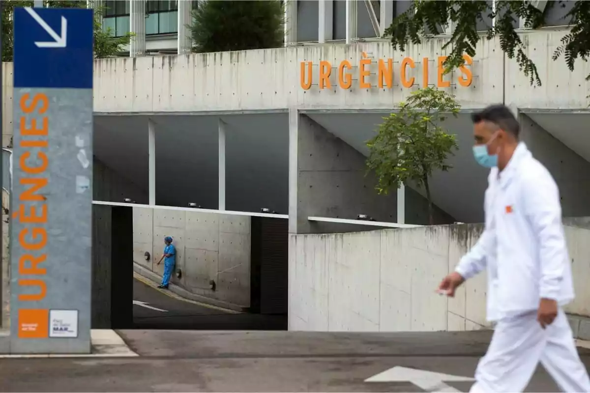 Entrada d’urgències d’un hospital amb un treballador de la salut amb bata blanca caminant i una altra persona amb uniforme blau recolzada a la paret