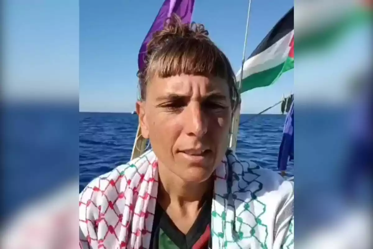 Persona con cabello recogido y flequillo, usando un pañuelo con diseño de cuadros, está en un barco sobre el mar con banderas de fondo y cielo despejado.