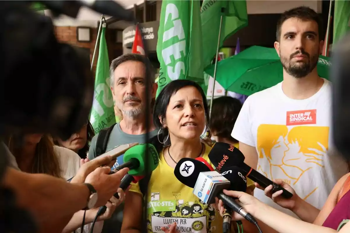 Una mujer rodeada de micrófonos da declaraciones a la prensa durante una manifestación, acompañada por otras personas y banderas verdes de un sindicato al fondo. Una mujer rodeada de micrófonos da declaraciones a la prensa durante una manifestación, acompañada por otras personas y banderas verdes de un sindicato al fondo.