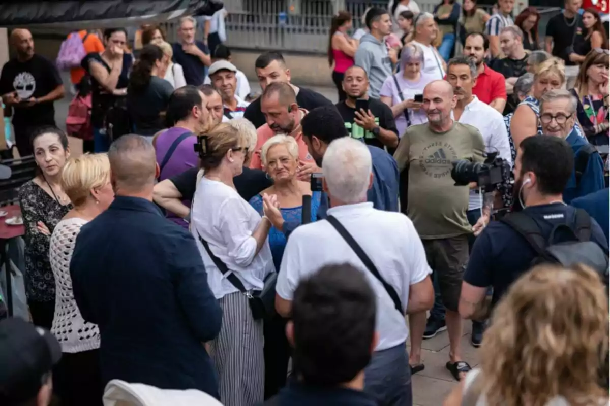 Un grup nombrós de persones es reuneix al carrer mentre algunes conversen i d’altres observen, amb un càmera gravant l’escena.