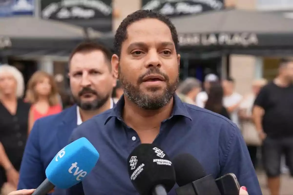 Un home amb barba i camisa blava parla davant de diversos micròfons mentre altres persones romanen al fons en un espai a l’aire lliure. Un home amb barba i camisa blava parla davant de diversos micròfons mentre altres persones romanen al fons en un espai a l’aire lliure.