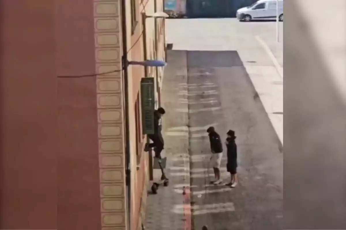 Tres persones observen un jove que intenta enfilar-se per la paret d’un edifici fent servir un patinet elèctric com a suport.