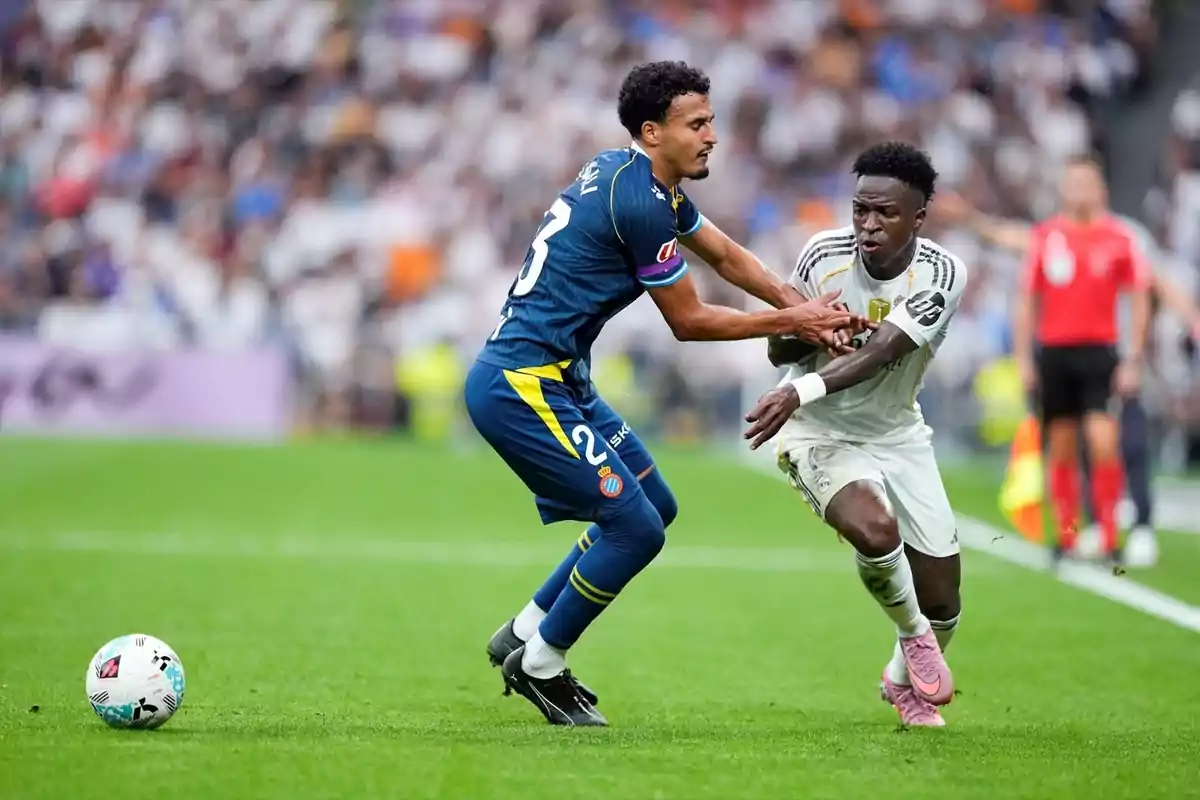 Dos futbolistes disputen la pilota durant un partit en un estadi ple d’espectadors