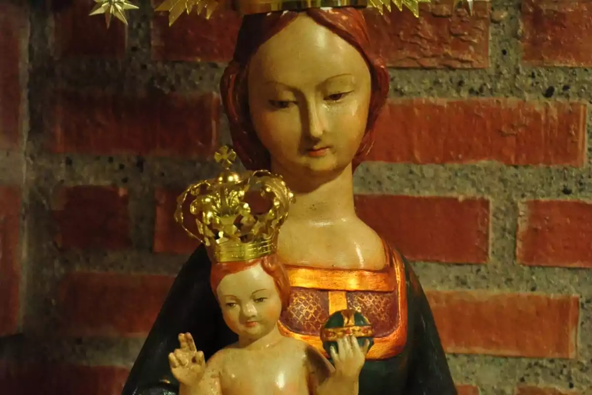 Escultura religiosa de la Virgen María con el Niño Jesús ambos con coronas doradas frente a una pared de ladrillos Escultura religiosa de la Virgen María con el Niño Jesús ambos con coronas doradas frente a una pared de ladrillos