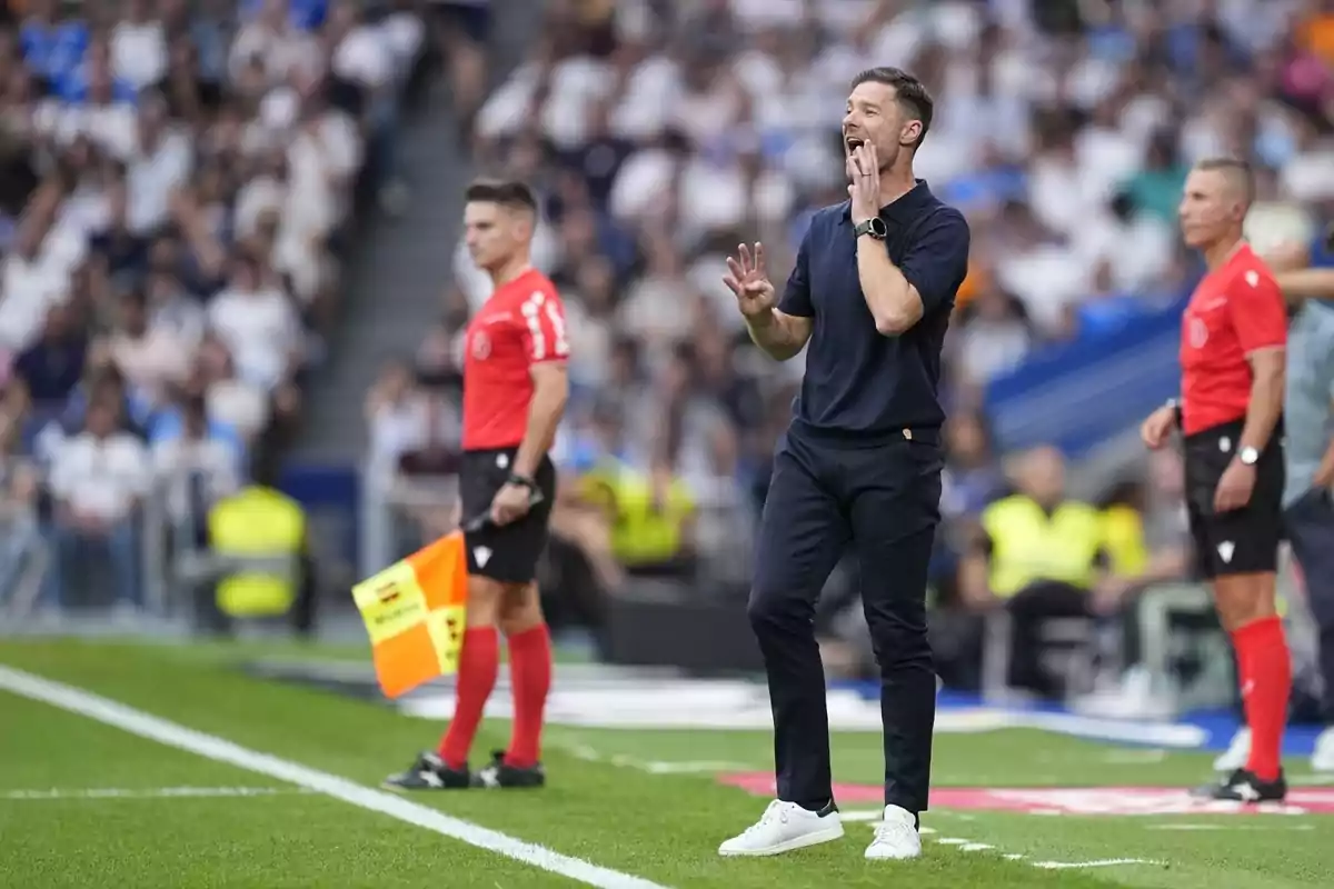 Entrenador de futbol donant indicacions a la banda durant un partit amb dos àrbitres assistents a prop i públic a les grades de fons.