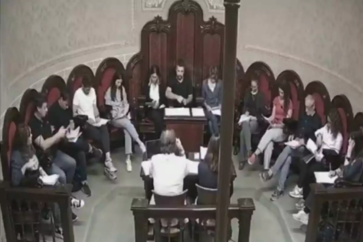 Personas sentadas en círculo en una sala con sillas de madera y respaldos altos, algunas revisando documentos y otras conversando