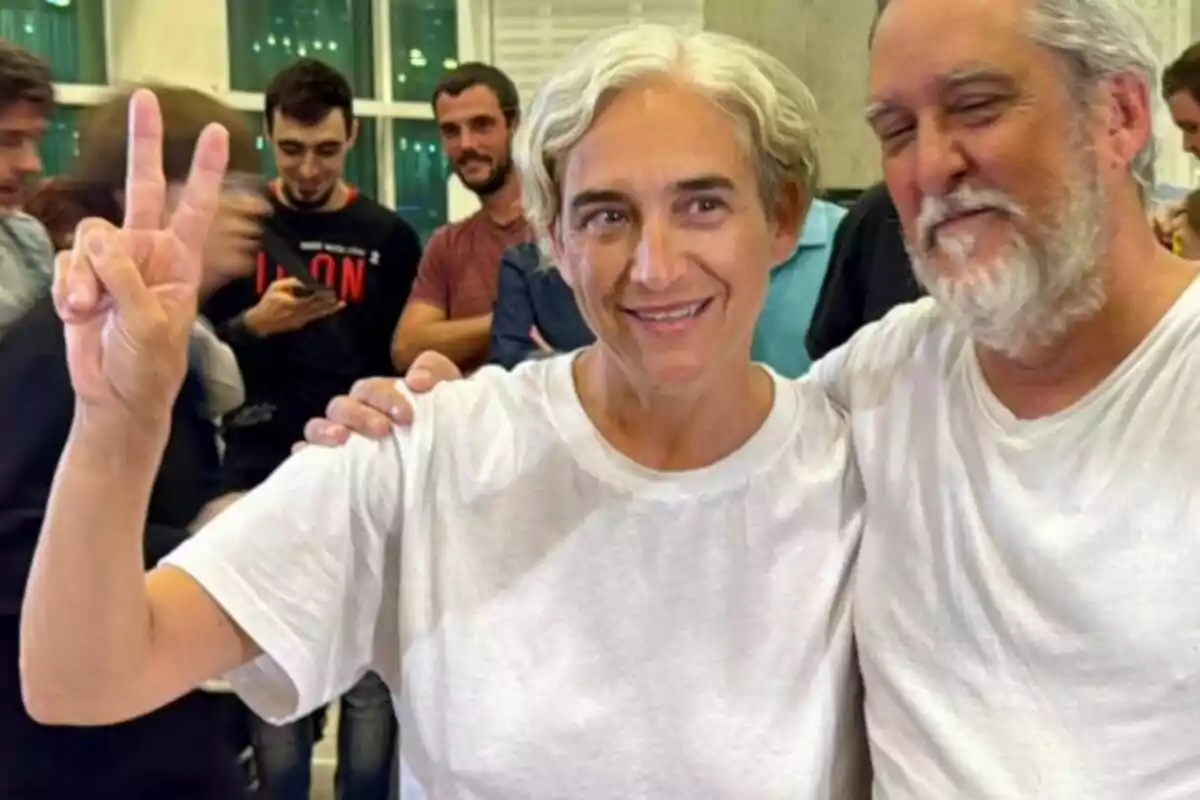 Dos personas sonríen y posan juntas, una de ellas hace el signo de la victoria con la mano, mientras varias personas se ven al fondo en un ambiente interior.