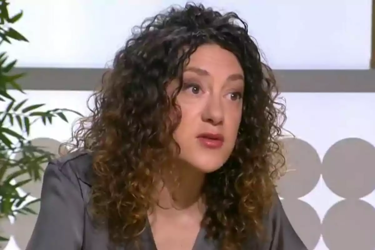 Mujer de cabello rizado y oscuro con expresión seria sentada en un estudio de televisión