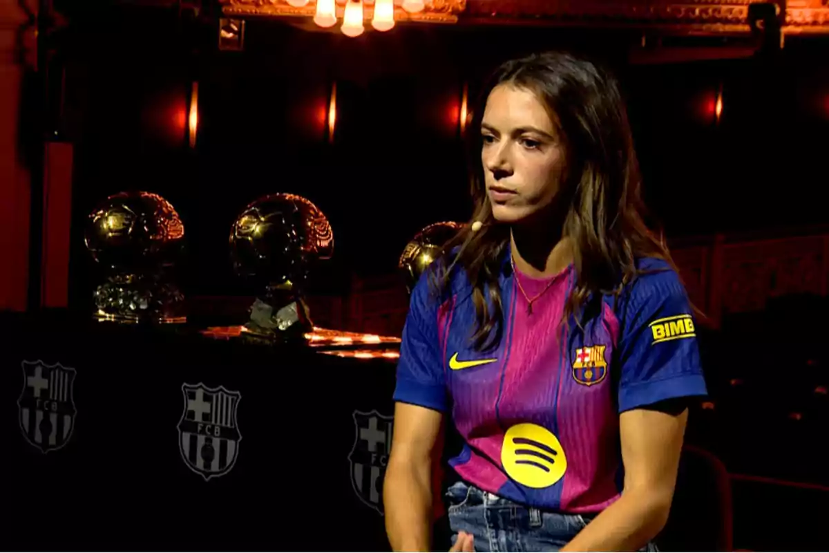 Futbolista amb samarreta del FC Barcelona asseguda al costat de tres trofeus Pilota d'Or en un ambient fosc i elegant