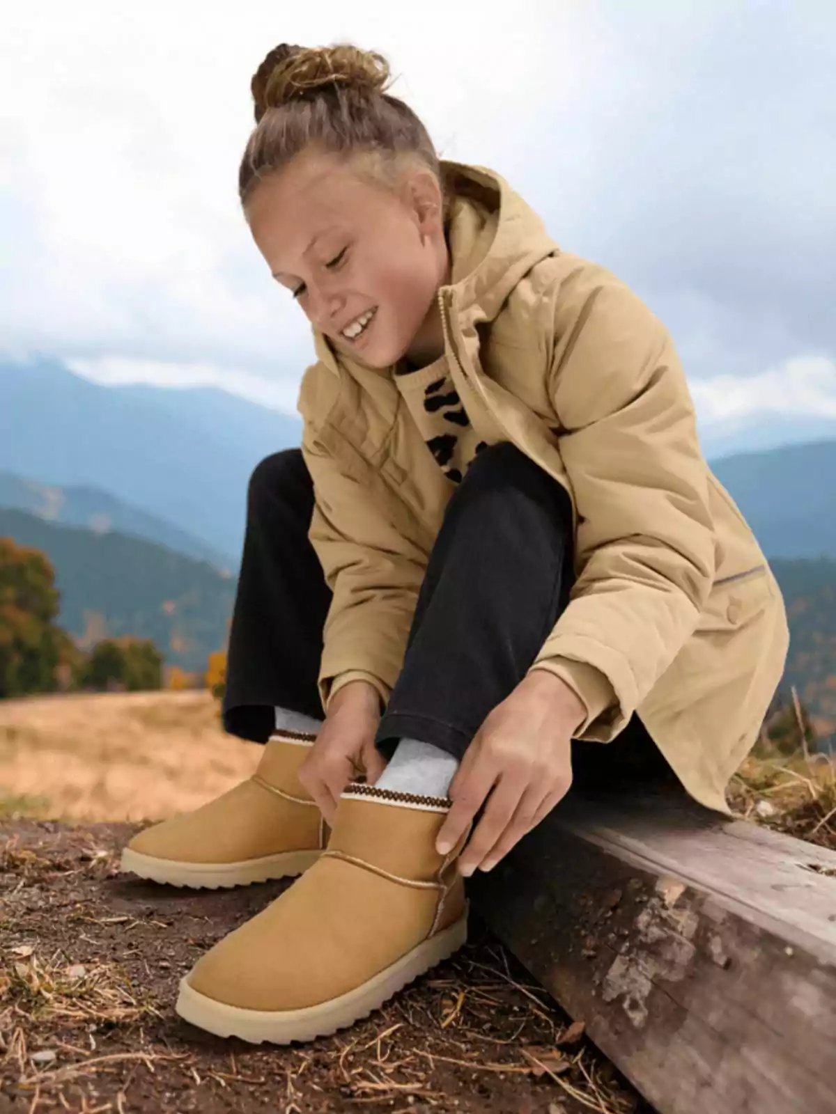 Niña sonriente sentada al aire libre en la montaña ajustándose unas botas color camel mientras usa una chaqueta beige y pantalones oscuros Niña sonriente sentada al aire libre en la montaña ajustándose unas botas color camel mientras usa una chaqueta beige y pantalones oscuros