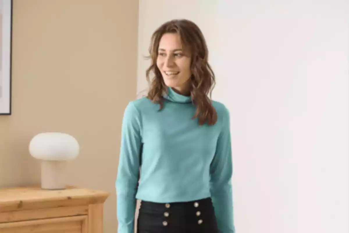 Mujer joven de cabello castaño sonriente con suéter azul claro y pantalón negro de botones en una habitación iluminada