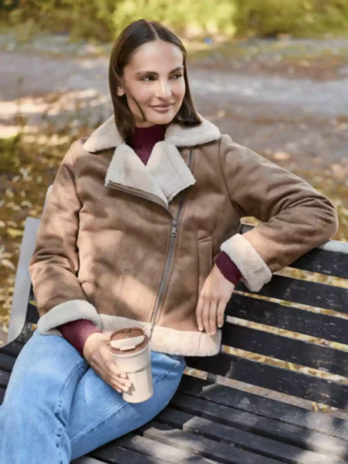Mujer sentada en una banca de parque con chaqueta marrón y taza térmica en la mano disfrutando de un día al aire libre Mujer sentada en una banca de parque con chaqueta marrón y taza térmica en la mano disfrutando de un día al aire libre