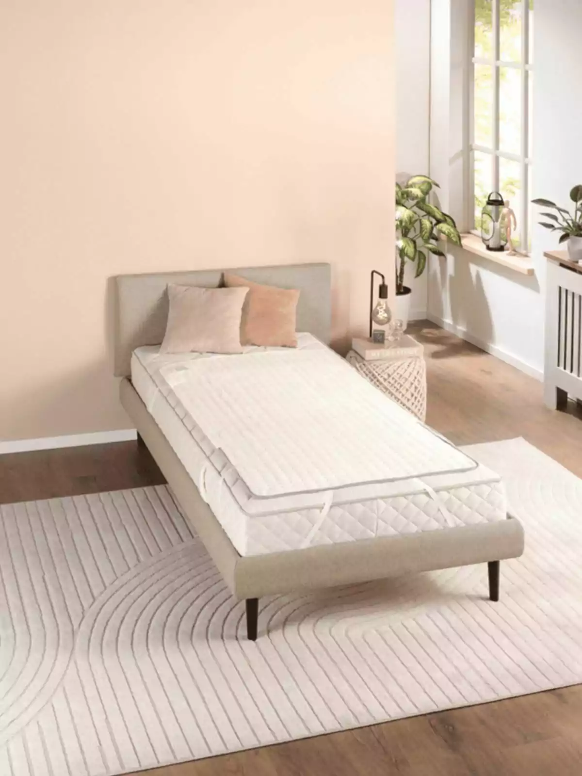 Cama individual con cabecera gris claro, dos cojines beige, sobre una alfombra blanca en una habitación luminosa con plantas junto a la ventana