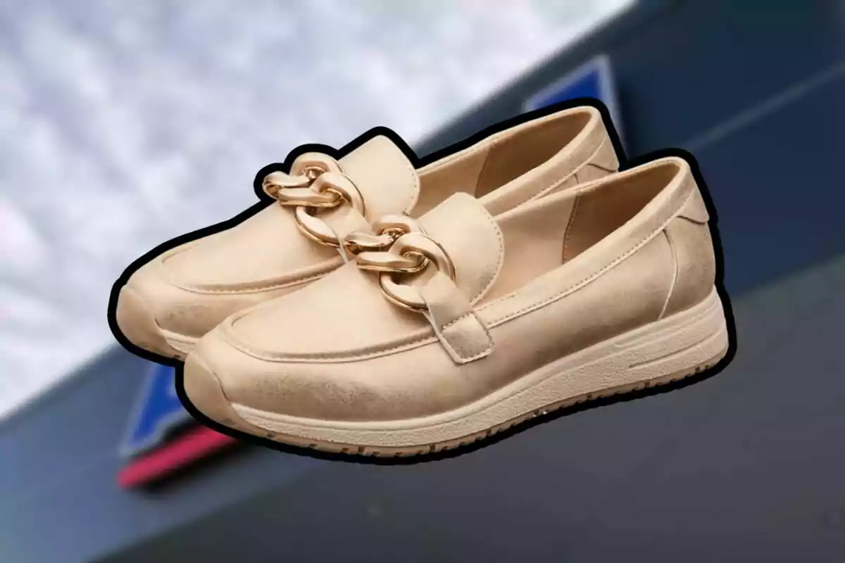 Sabates tipus mocassí de color beix amb detall de cadena daurada sobre un fons desenfocat. Sabates tipus mocassí de color beix amb detall de cadena daurada sobre un fons desenfocat.