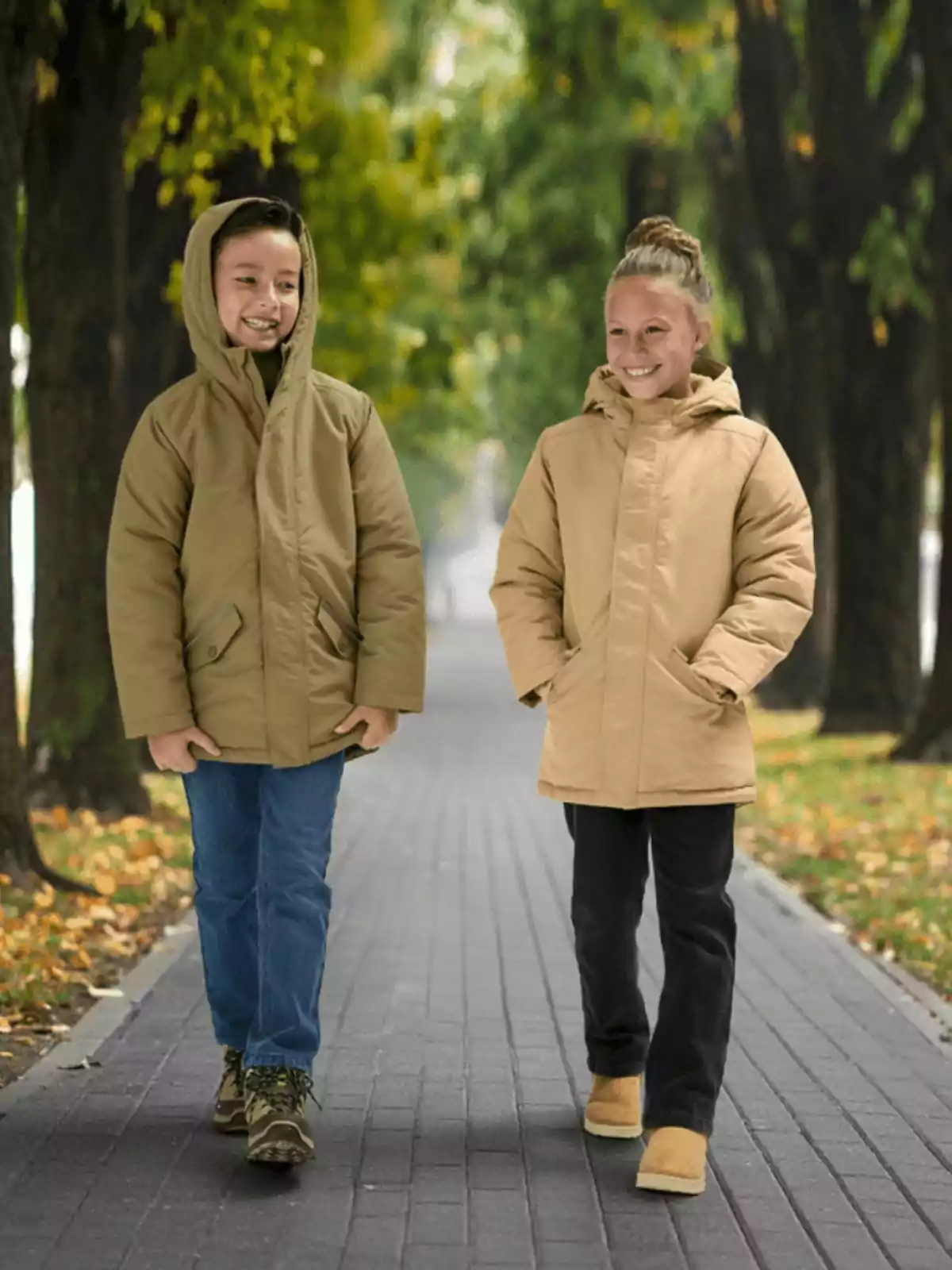 Dos niños sonrientes caminan por un sendero rodeado de árboles vistiendo abrigos beige y pantalones largos