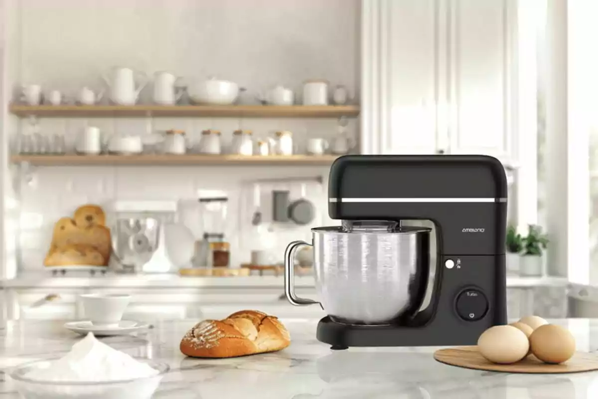 Batedora de cuina negra sobre una tauleta de marbre amb ous, pa i un bol de farina en una cuina moderna i lluminosa