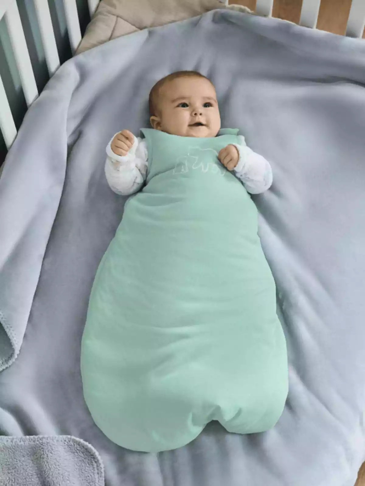 Bebé acostado en una cuna sobre una manta gris usando un saco de dormir verde claro Bebé acostado en una cuna sobre una manta gris usando un saco de dormir verde claro