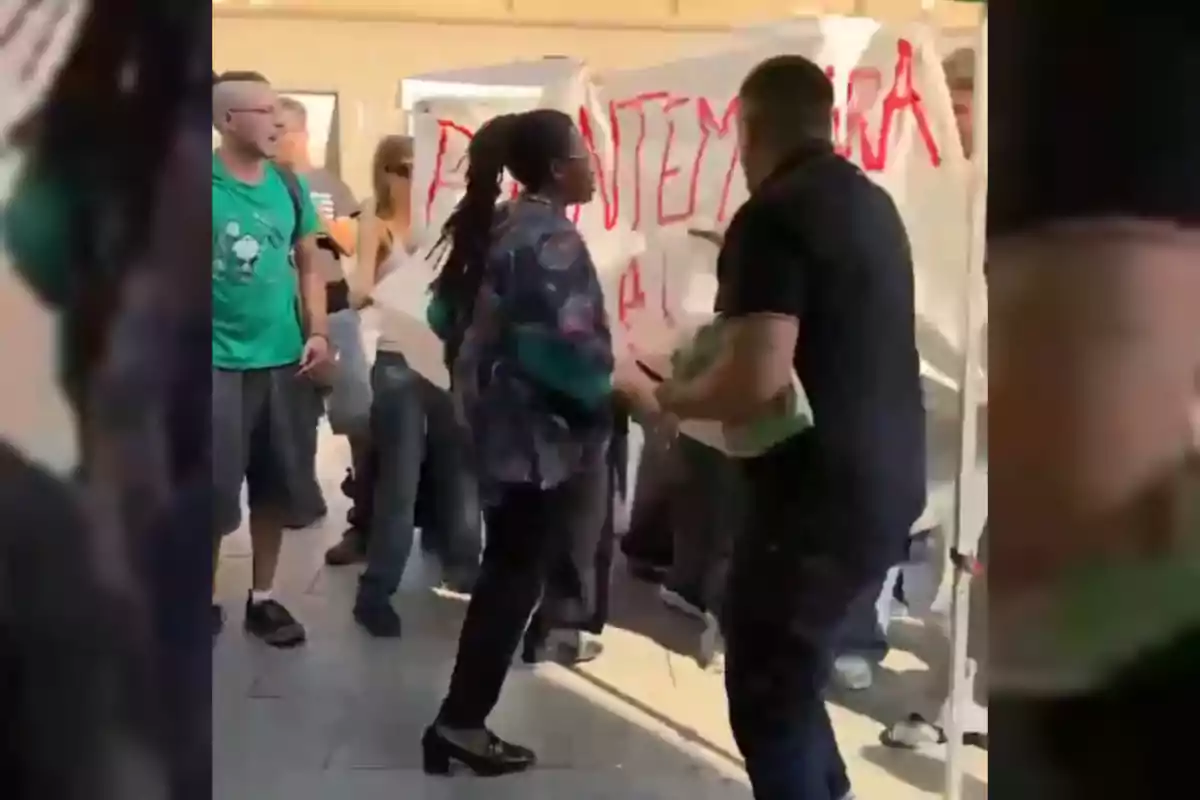 Diverses persones discuteixen davant d’una manta blanca amb lletres vermelles en el que sembla ser una manifestació al carrer.