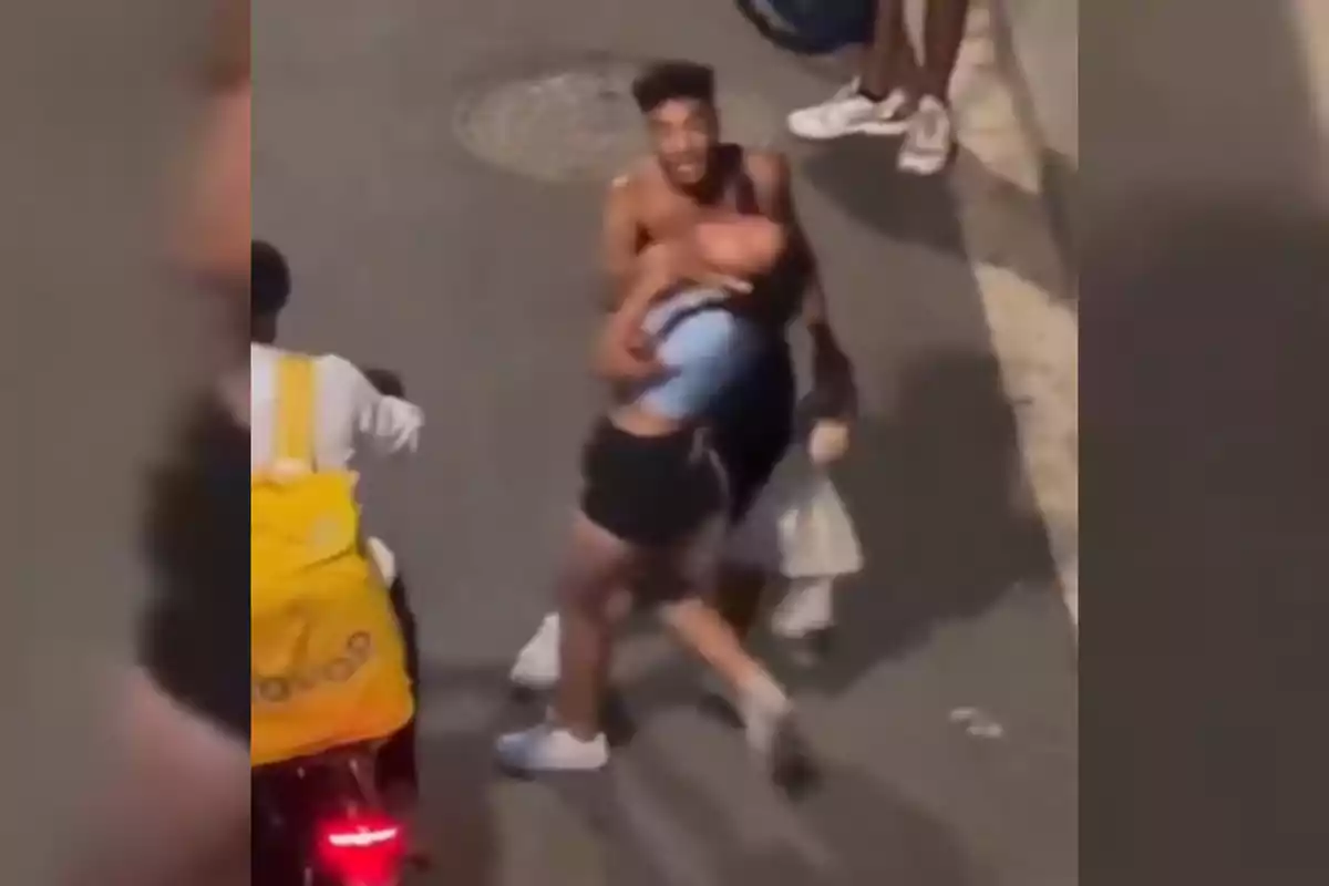 Un hombre sin camiseta sostiene a una mujer por el cuello en una calle mientras una persona con mochila amarilla pasa en bicicleta cerca de ellos