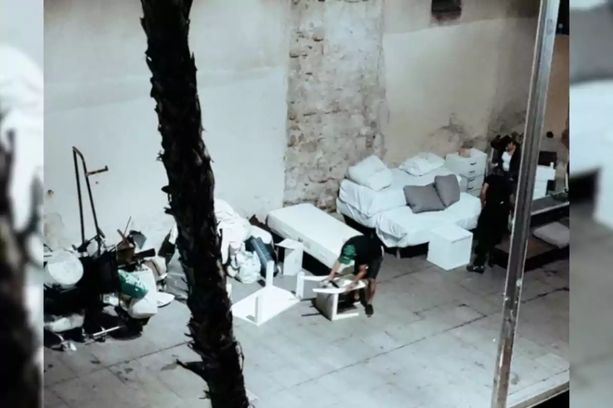 Personas armando muebles y organizando colchones y objetos en una calle junto a una pared de piedra