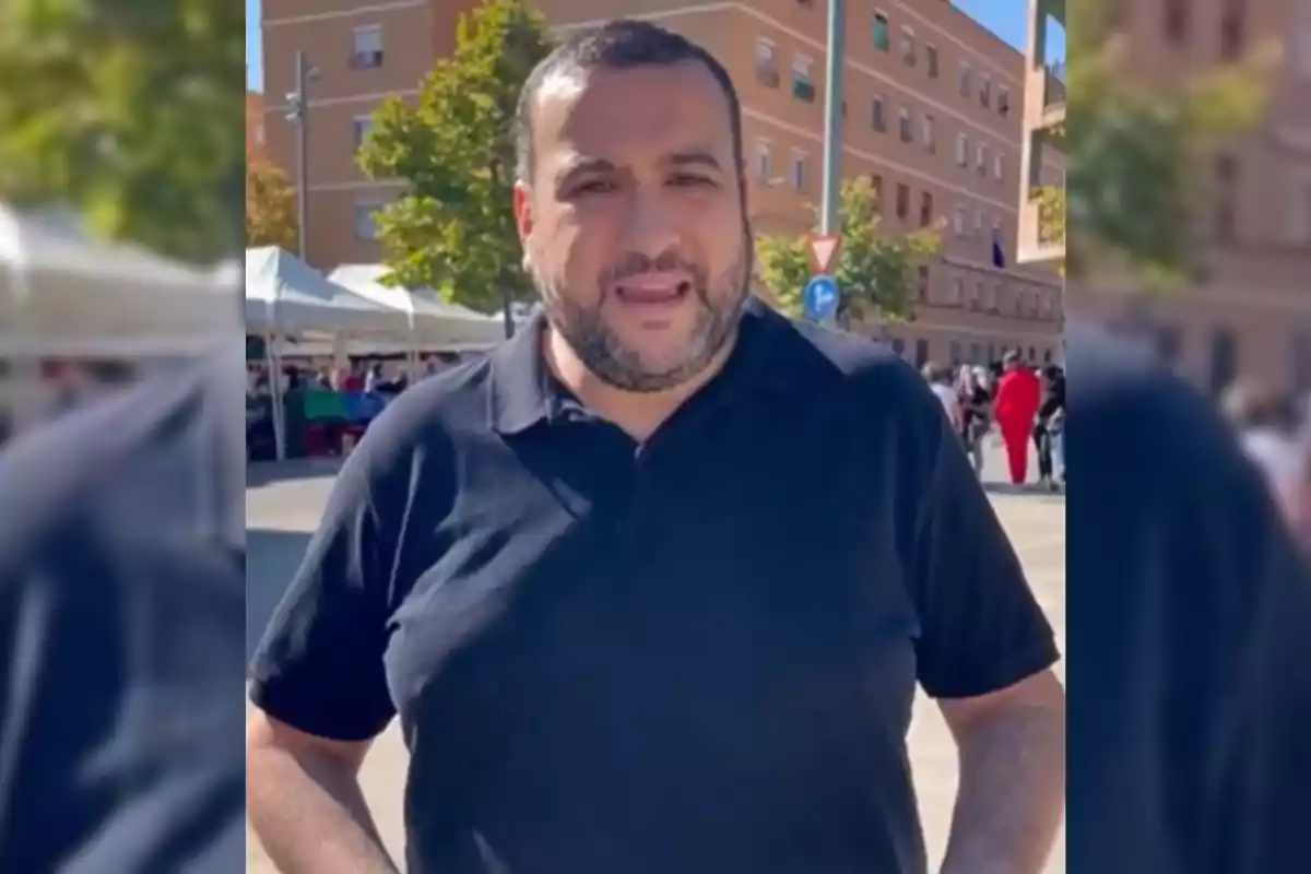 Hombre de barba con camiseta negra de pie al aire libre en una plaza con edificios y árboles al fondo