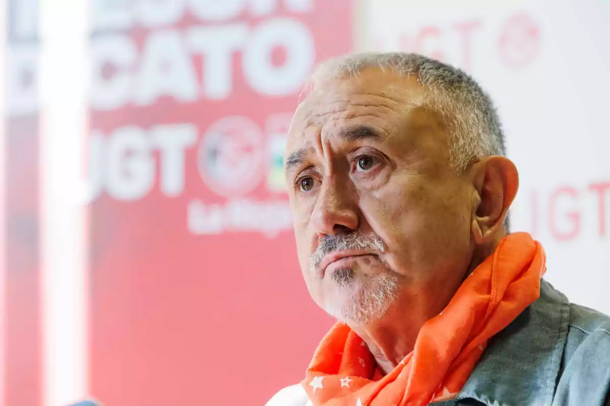 Hombre mayor con cabello canoso y barba, usando un pañuelo naranja con estrellas, frente a un fondo rojo con el logo de UGT desenfocado.