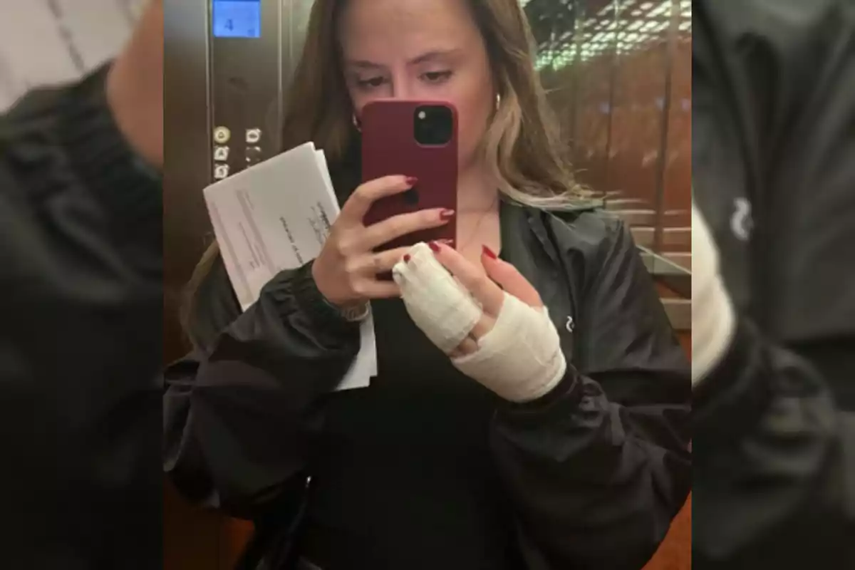 Mujer tomándose una selfie en un elevador con la mano vendada y sosteniendo unos papeles