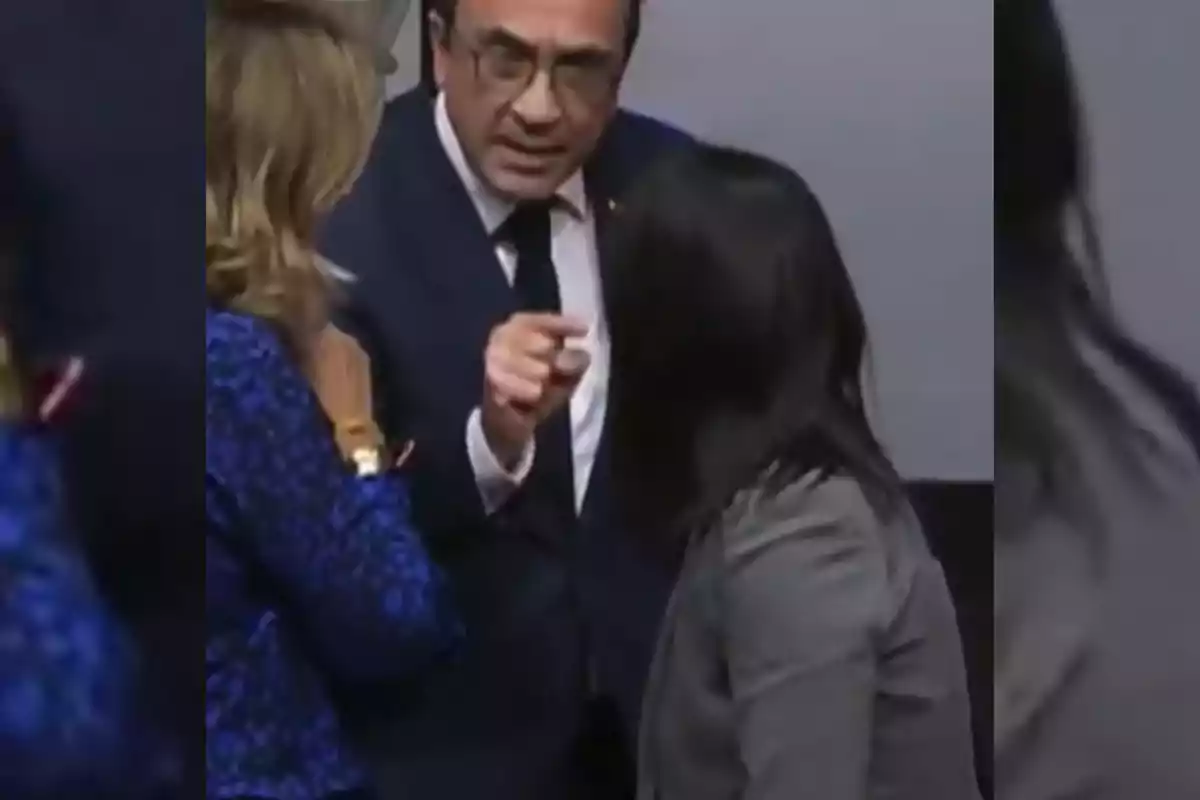 Un hombre con gafas y traje oscuro señala con el dedo mientras habla con dos mujeres en un entorno formal.