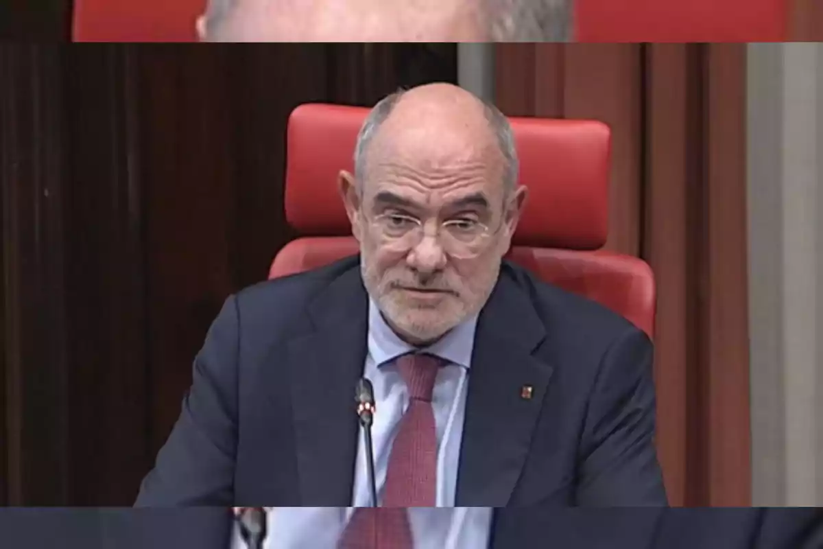 Hombre mayor con gafas y barba canosa sentado en una silla roja durante una reunión formal