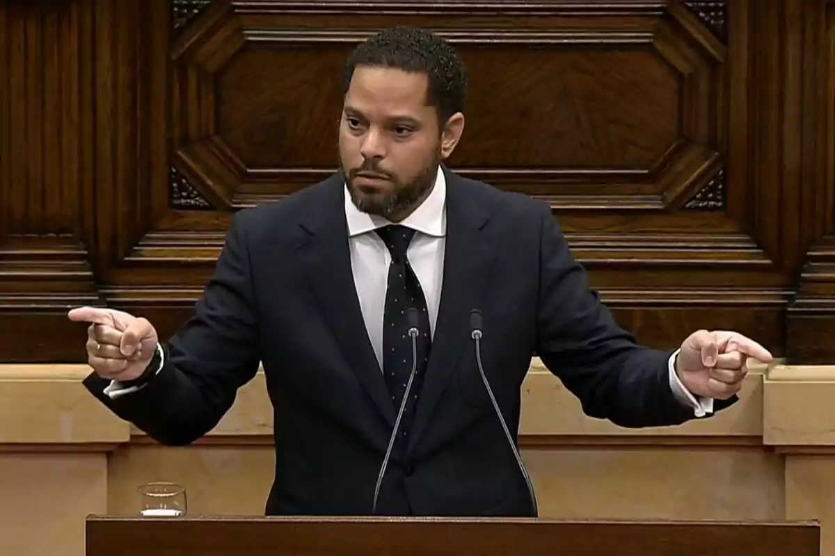 Hombre de traje oscuro hablando en un podio de madera y señalando hacia ambos lados con las manos