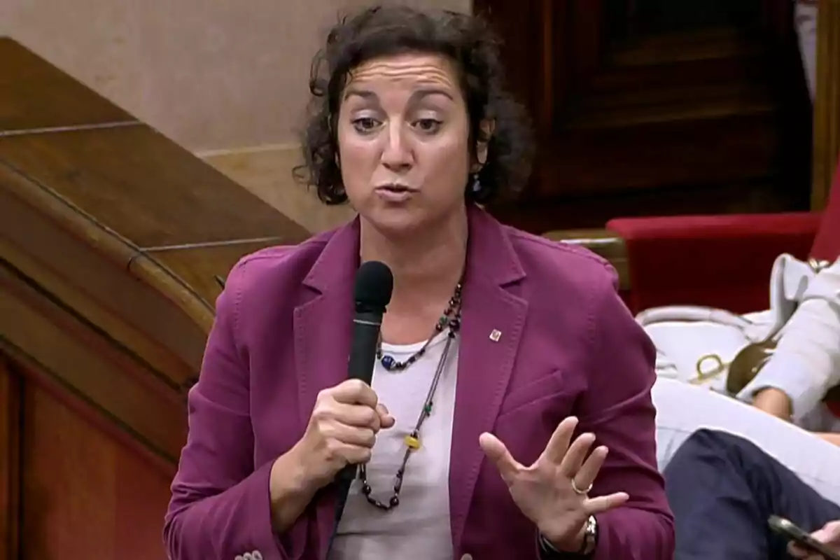 Mujer de cabello corto y rizado con chaqueta fucsia hablando mientras sostiene un micrófono en un entorno tipo parlamento Mujer de cabello corto y rizado con chaqueta fucsia hablando mientras sostiene un micrófono en un entorno tipo parlamento