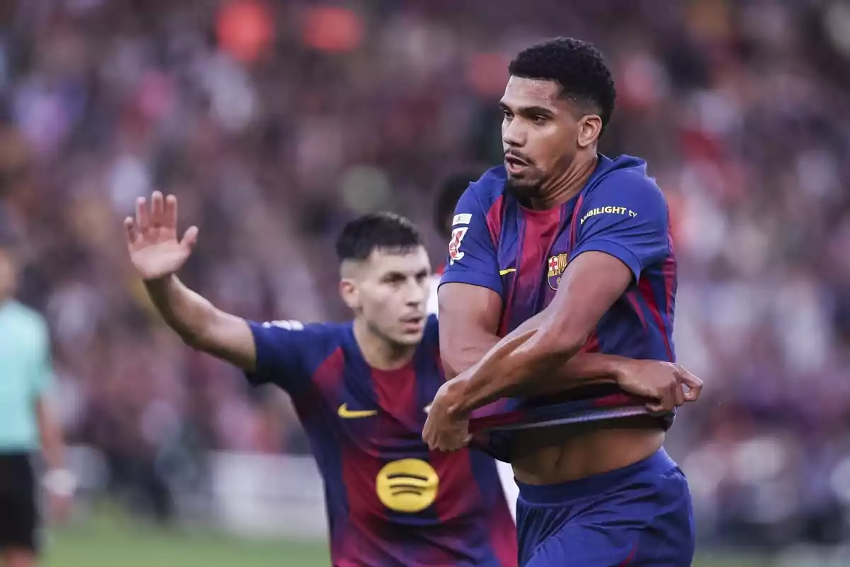 Dos futbolistes del Barcelona amb l’uniforme blaugrana durant un partit, un d’ells aixecant la samarreta i l’altre amb la mà aixecada al fons del camp.