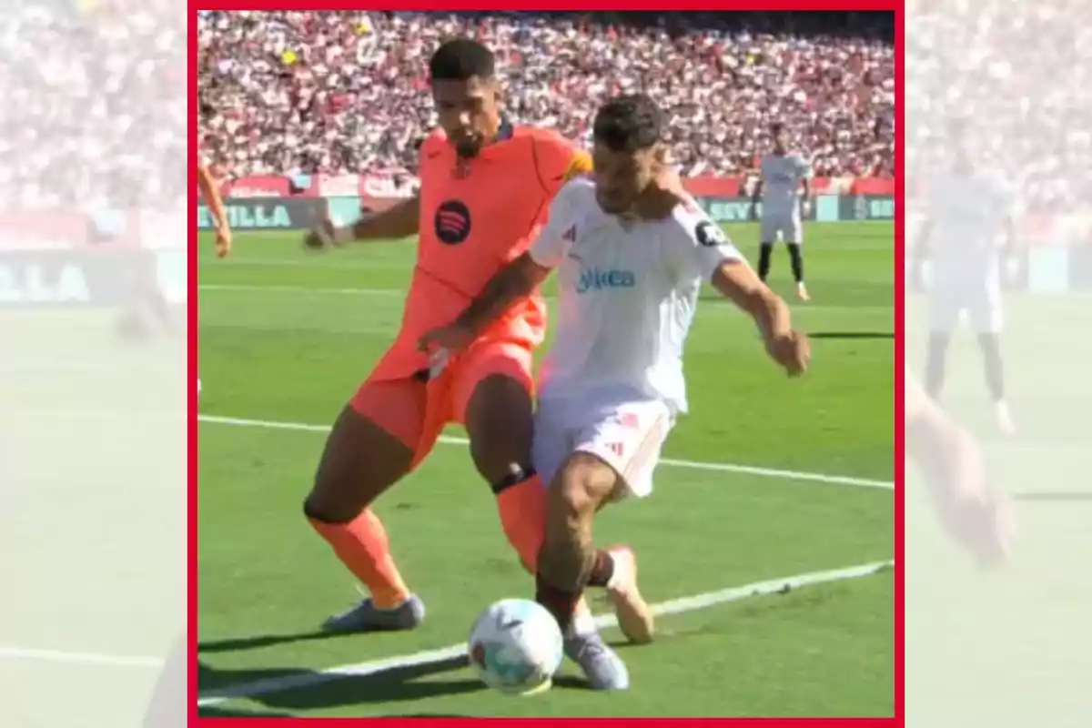 Dos futbolistas disputan el balón cerca de la línea de banda durante un partido, uno con uniforme naranja y otro con uniforme blanco, mientras el público observa desde las gradas.